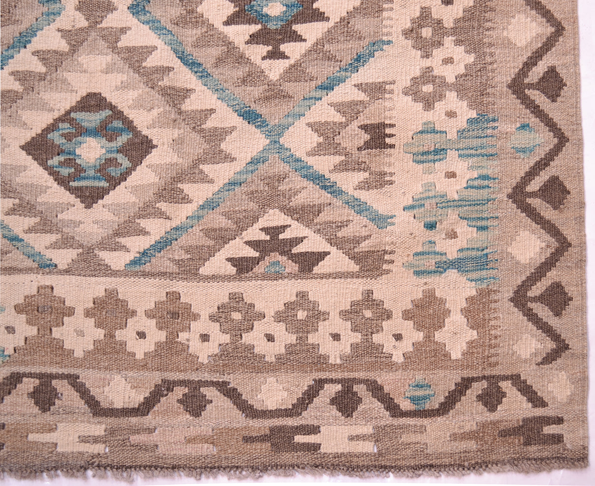 A close-up of a woven kilim rug featuring a geometric pattern in beige, brown, and teal. The rug shows detailed texture and the corner of its border with small fringe visible at the bottom edge.