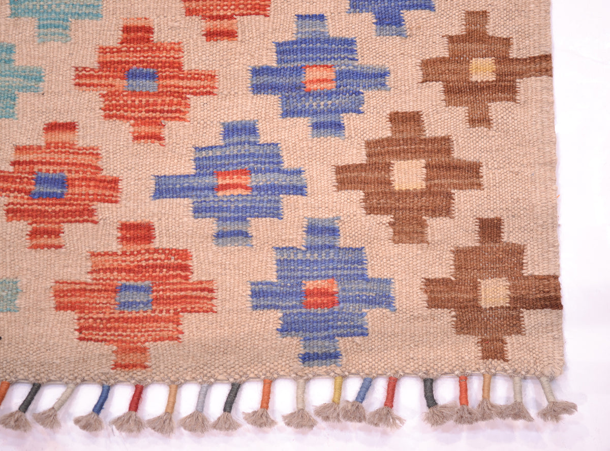 A close-up of a woven Afghan Kilim long runner with a beige base and geometric motifs in rust red, medium blue, and brown. The bottom edge has multicoloured wrapped tassels.