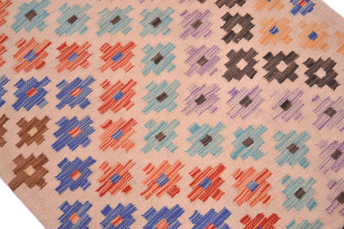 A close-up of a woven Kilim long runner with a repeating geometric ziggurat pattern in blue, rust red, light blue, purple, brown, and orange on a warm beige background.