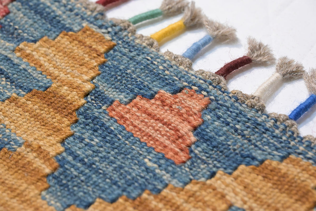 Close-up of a woven Giant Handwoven Afghan Kilim rug with a geometric pattern in blue, tan, and terracotta, finished with multicolored tassels on a white background.