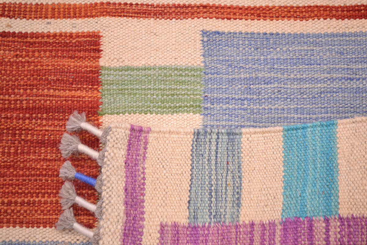 Close-up of the handwoven back corner of an Afghan modern Kilim runner rug with a multicolour geometric pattern including orange, beige, blue, green, purple, and turquoise blocks, finished with grey tassels.