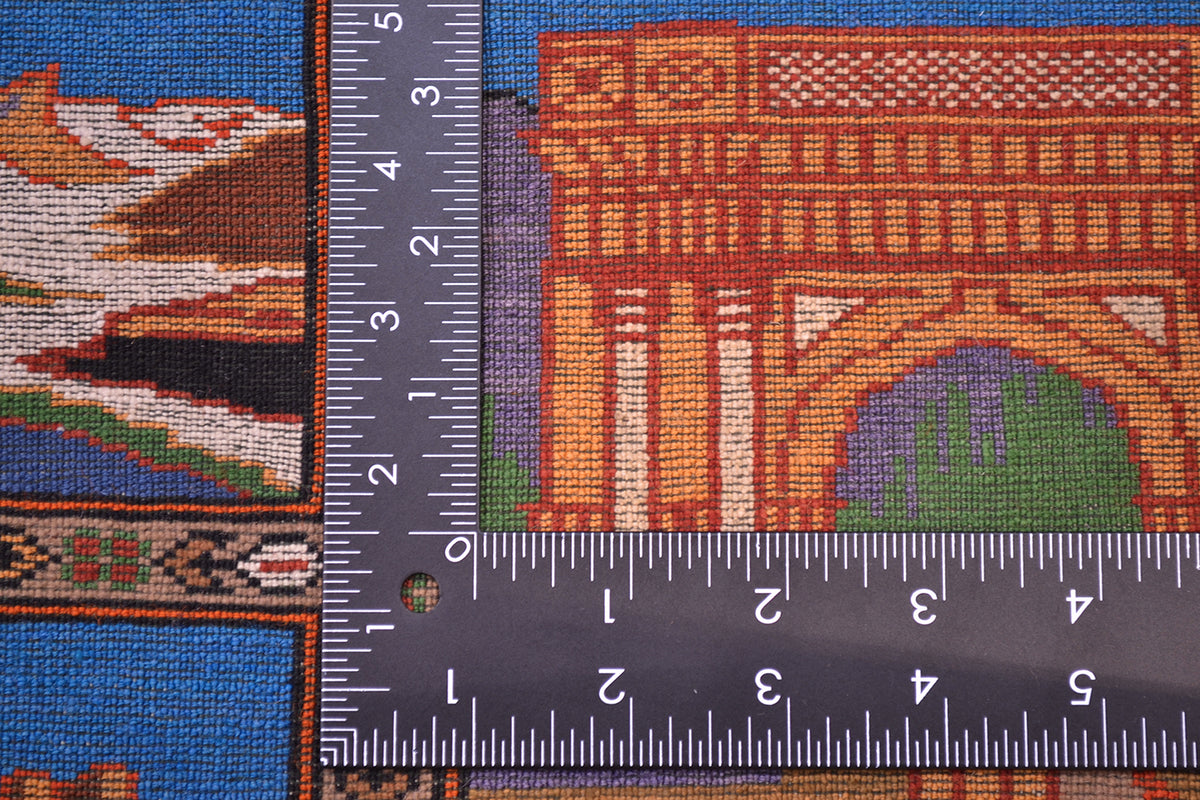 Close-up of a hand-knotted fine Pictorial Afghan rug with a dark measuring ruler placed over it to show scale of the knot density, featuring an orange archway motif and blue border areas.