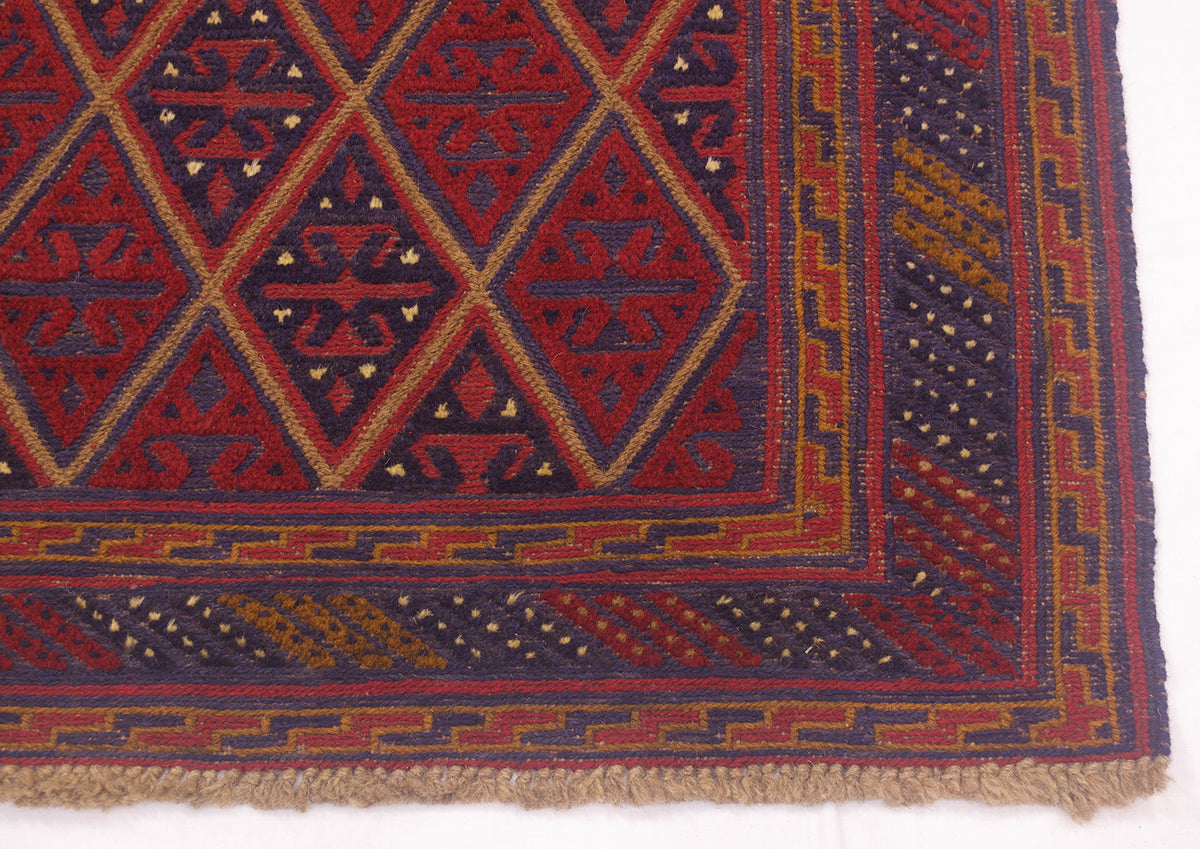 A close-up of a patterned Mushwani rug corner showing a geometric diamond design in red, navy, and tan with a fringed edge on a white background.