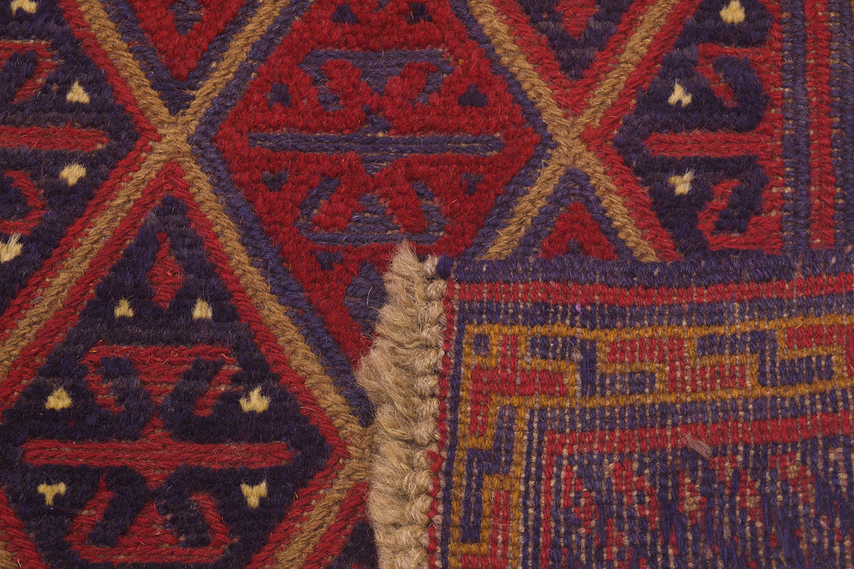 Close-up of the weave of a patterned handmade Mushwani square rug showing a geometric design in red, navy blue, and tan, with a frayed woven edge visible.