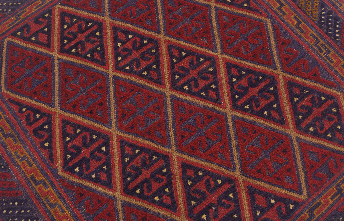 A close-up of a woven Mushwani square wool rug with a geometric diamond pattern in deep red, navy blue, and tan.