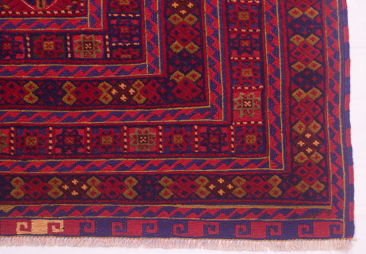 A close-up of a fine Afghan rug corner showing deep red, navy blue, and tan geometric motifs with white fringe along the bottom edge.