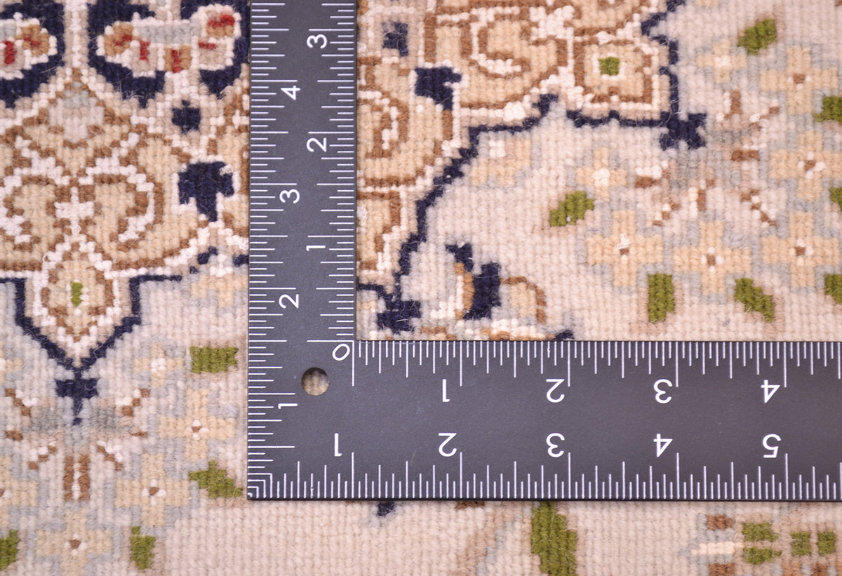 Close-up of the hand-knotted back of an Indo Persian Nain rug with a metal L-square ruler placed at the corner to show the scale of the design. The rug has a cream base with navy blue, brown, and muted green geometric and floral motifs.
