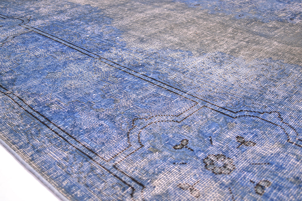 Close up detail of blue and grey brown vintage overdyed carpet with geometric detailing outlining in dark black charcoal. Exposed cttonweft and warps and shaved distressed pile in blues.