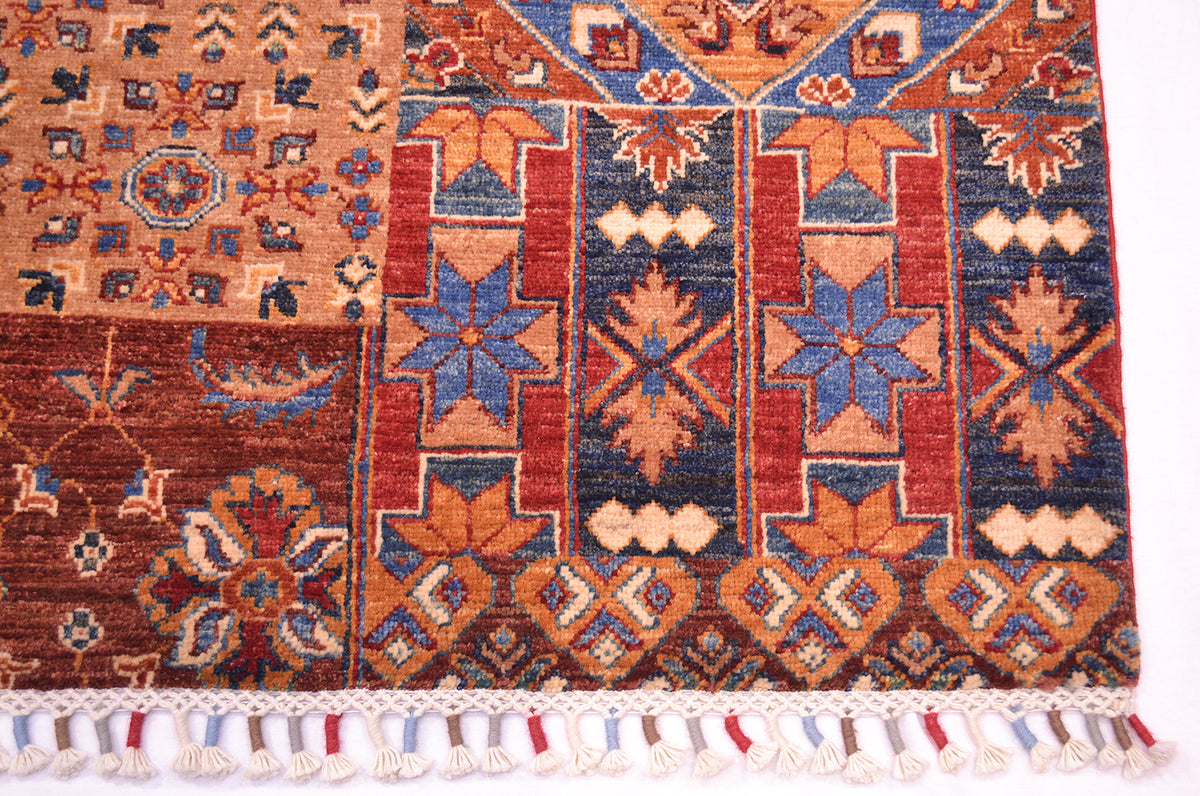 A close-up of a corner of a patterned Patchwork design rug with geometric and floral motifs in rust red, navy blue, tan, and cream, finished with a white tassel fringe.