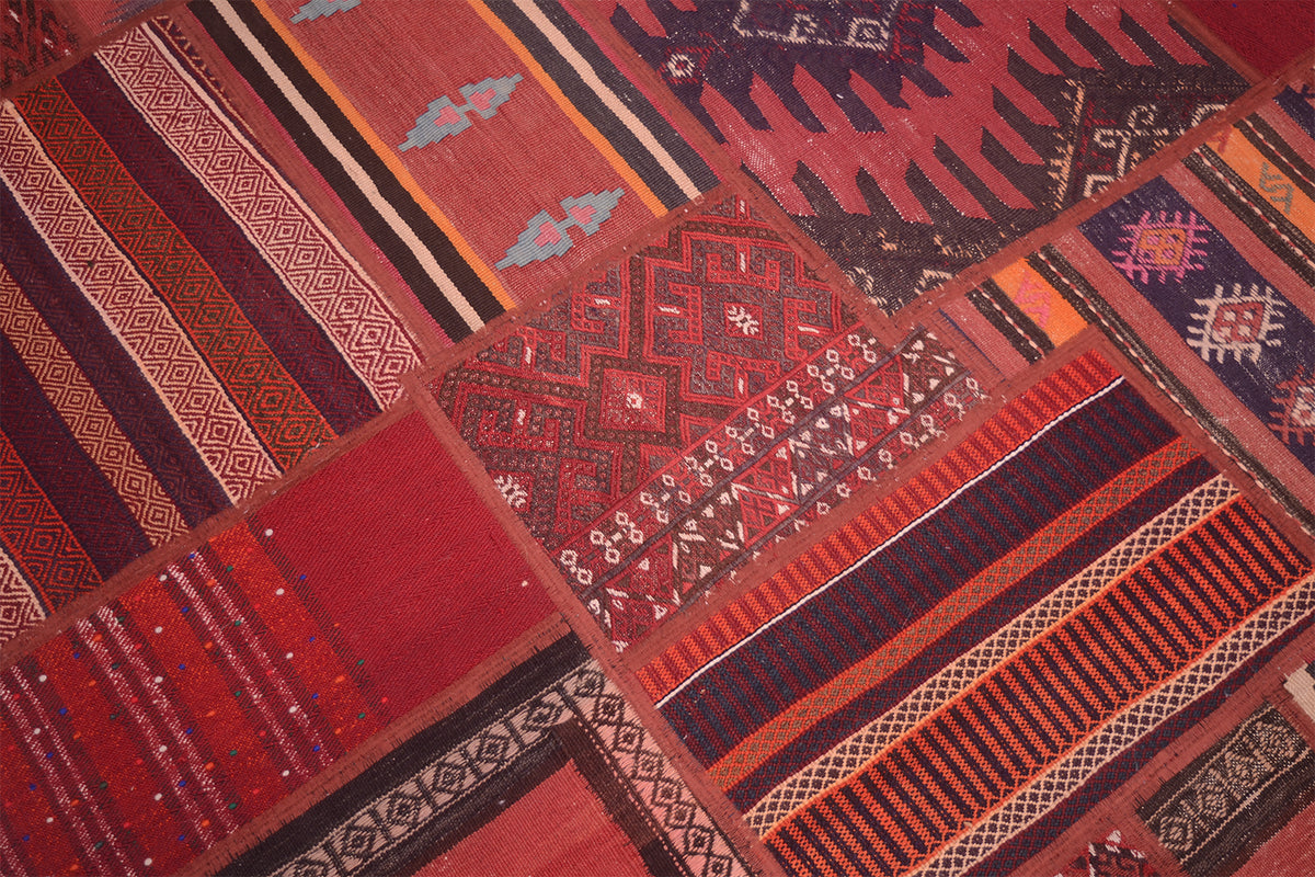 A close-up of a patchwork kilim rug made of multiple red, rust, orange, brown, and black patterned sections with geometric and striped motifs.