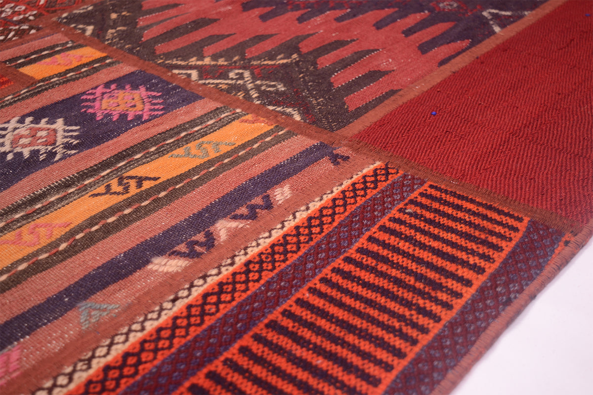 A close-up of a handmade Patchwork Kilim rug with geometric patterns in rust red, orange, brown, navy, and cream, laid diagonally across a white surface.