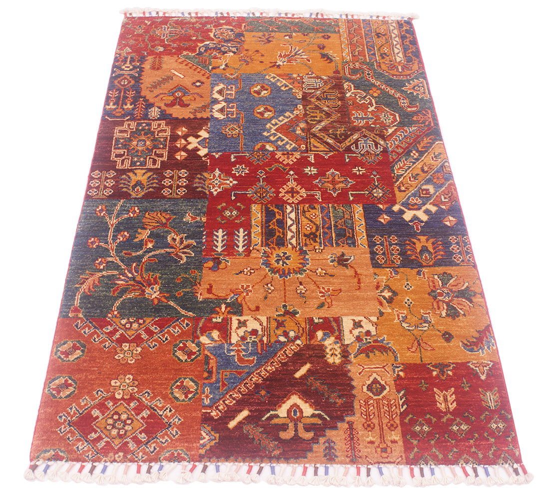 A rectangular Afghan patchwork-style rug with a traditional geometric and floral pattern, featuring multiple panels in rust red, terracotta, navy blue, ochre orange, and cream, finished with white fringe on the ends.