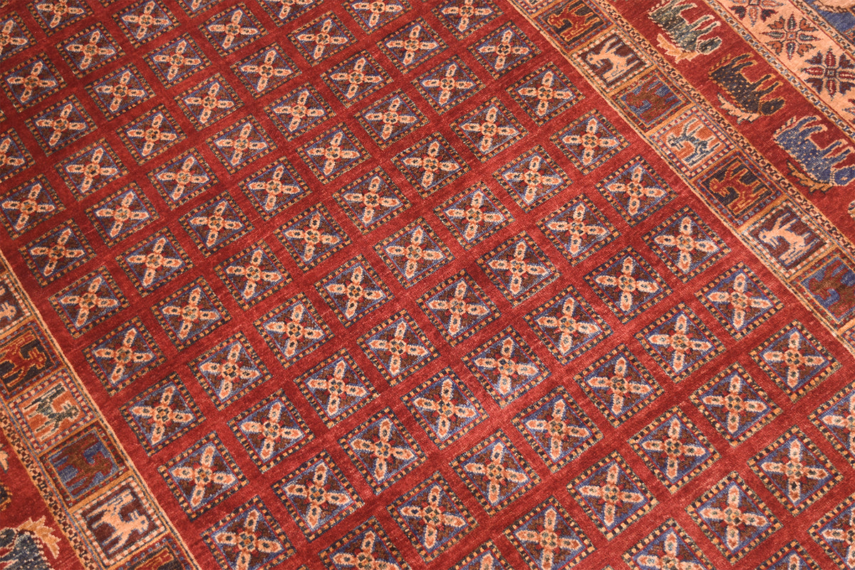 A close-up of a patterned Handmade Afghan Pazyryk carpet with a deep red field, repeating cream and blue geometric cross motifs, and a decorative border in beige, blue, and red.