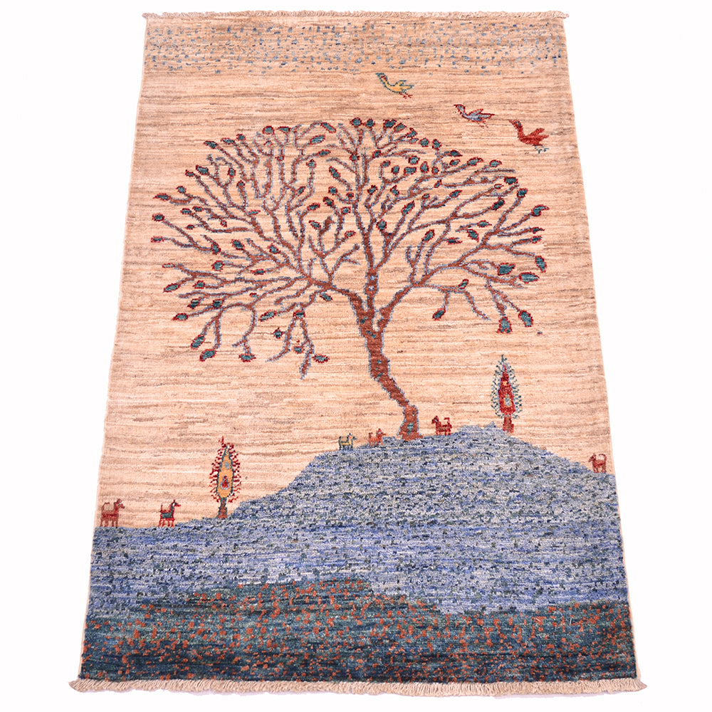 A rectangular handwoven pictorial Loribaft small rug featuring a stylized tree on a hill with small birds and animals, set against a light beige background with a blue textured lower section.