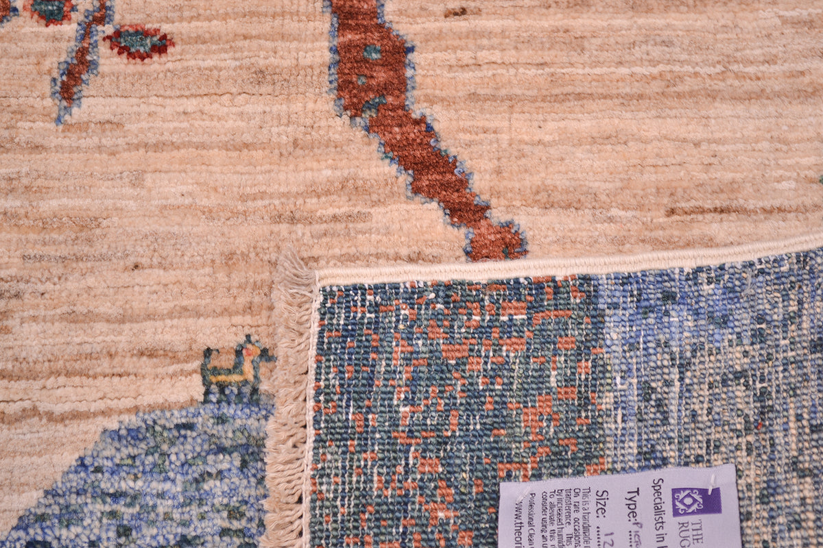 Close-up of a pictorial Loribaft small rug rug showing the corner turned up revealing the hand-knotted weave, fringed edge, and product tag. The rug features a light beige and tan base with blue, rust red, and grey woven patterns.