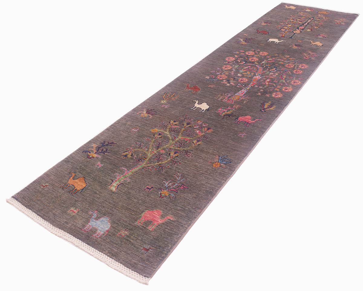 A long rectangular Pictorial grey Loribaft runner with a textured pewter background, featuring a whimsical folk-art style design of trees, flowers, and animals in muted pink, orange, blue, and cream tones. The rug has a light beige fringed edge and is photographed on a clean white background.
