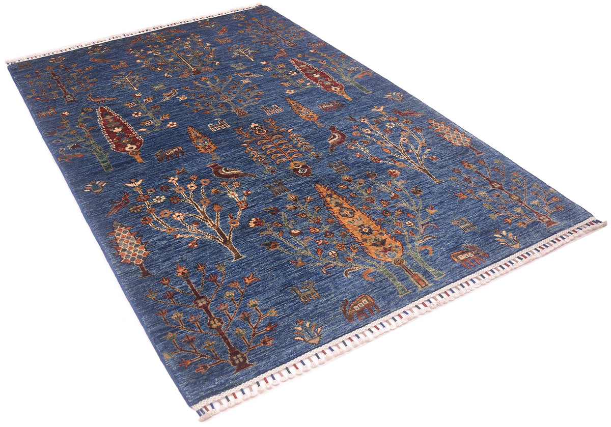 A rectangular pictorial blue Loribaft rug with a blue background and an intricate, traditional tribal-style pattern featuring trees, birds, and geometric shapes in brown, beige, and muted orange. The rug has a white fringed border on one end.