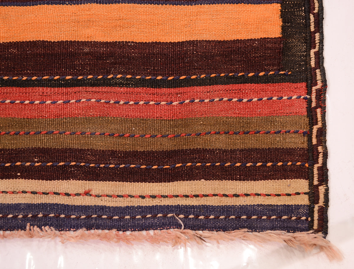 A close-up of a striped woven vintage Kilim runner rug corner showing horizontal bands in orange, dark brown, rust red, olive green, beige, and navy blue with fringed edges.