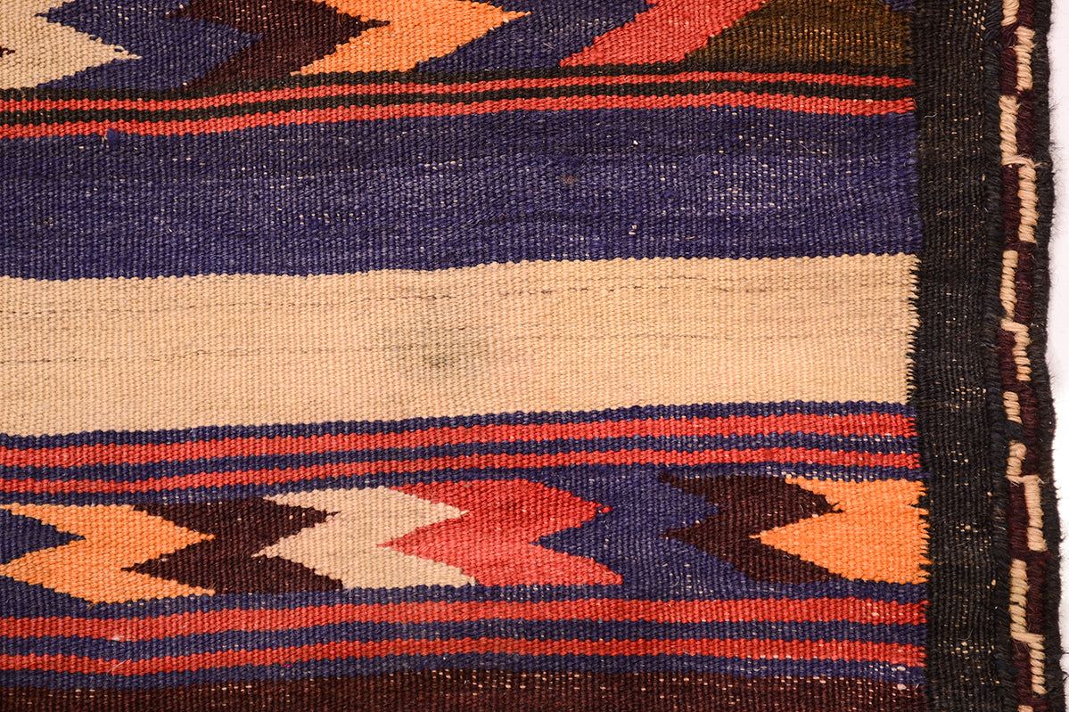 A close-up of a woven Kilim wide runner with horizontal bands and geometric chevron motifs in dark blue, cream, orange, red, and brown, with a dark edge binding visible on the right.