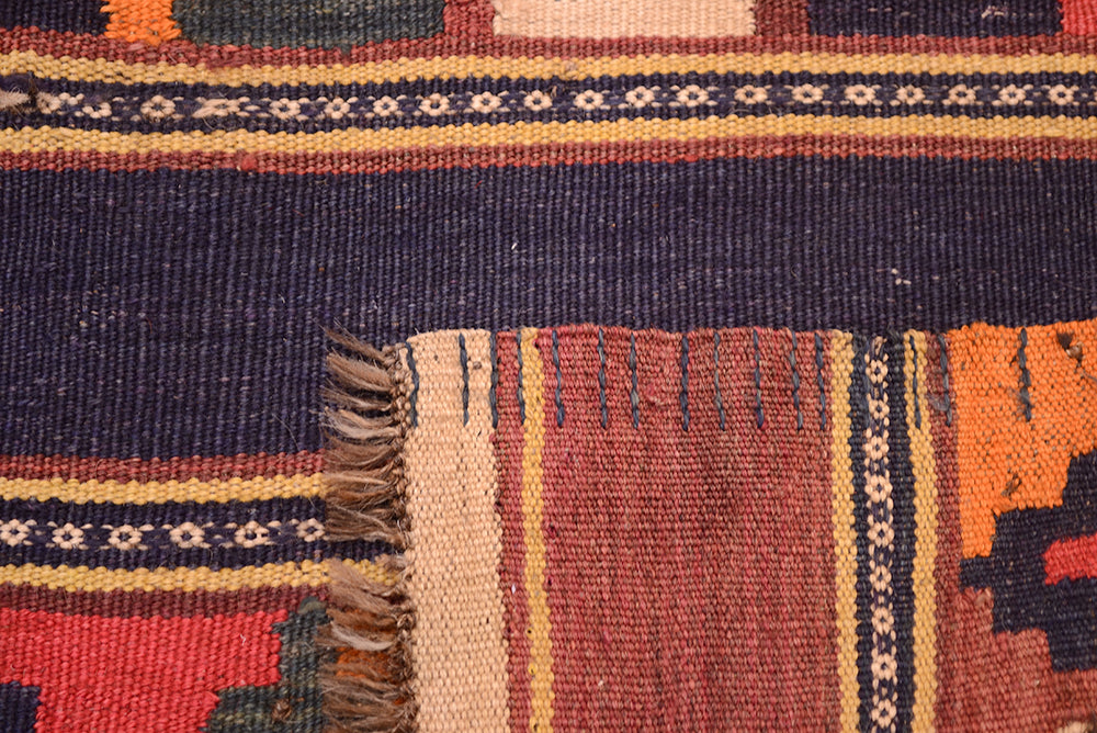Close-up of a corner underside of a hand woven old Afghan kilim runner rug showing a dark navy blue field, patterned borders in cream, rust, and orange, and a fringed edge.
