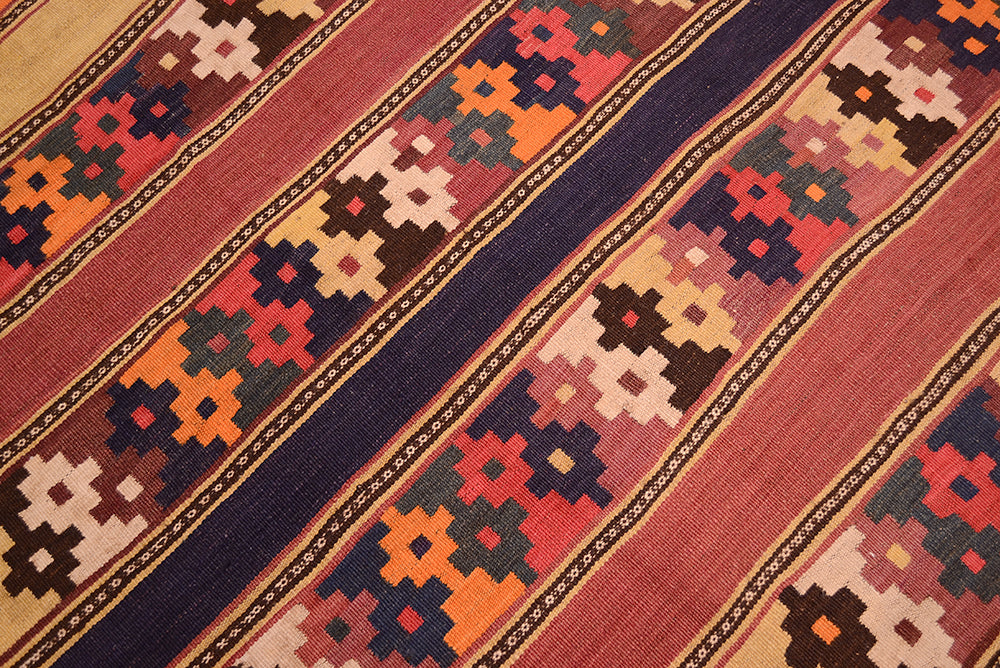 A close-up of a woven old Afghan kilim rug with diagonal stripes and geometric patterns in rust red, navy blue, orange, beige, and brown.