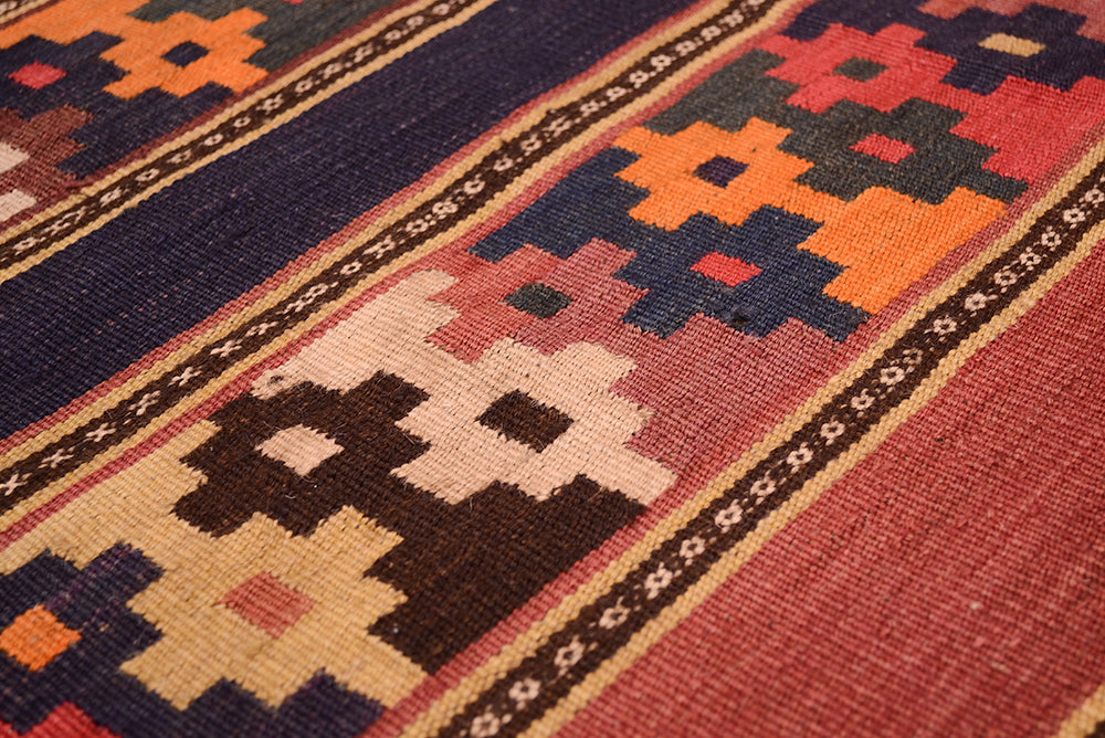 Close-up of a hand woven old Afghan Kilim runner rug with a geometric pattern in red, orange, beige, brown, and dark blue, showing detailed fabric texture.