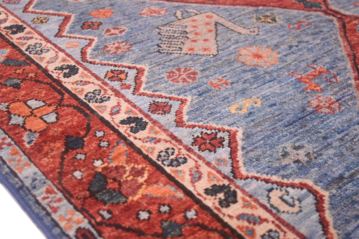 A close-up of a traditional patterned Afghan tribal Qashqai rug with a dusty blue field, a red and peach zigzag border, and dark floral motifs.