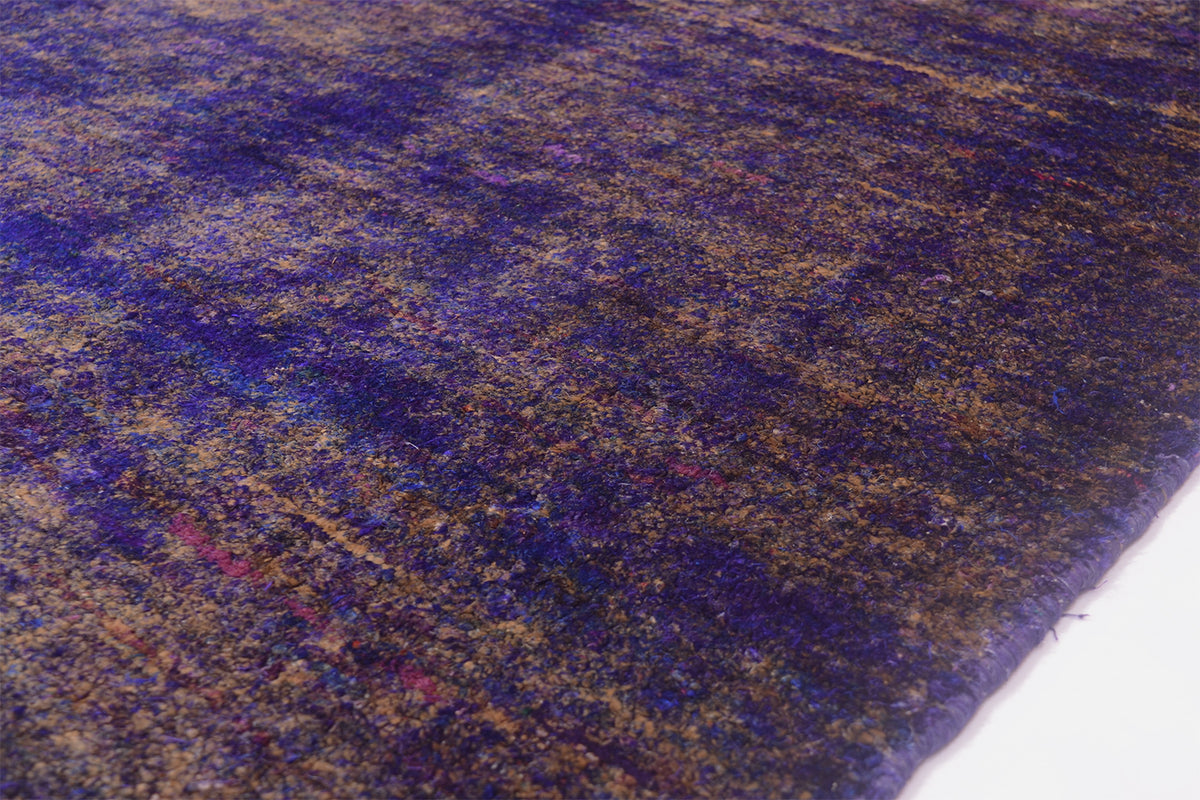 A close-up of a Handmade Sari Silk rug showing a mottled pattern of deep purple, blue, and gold tones, with a white surface visible at the bottom right corner.