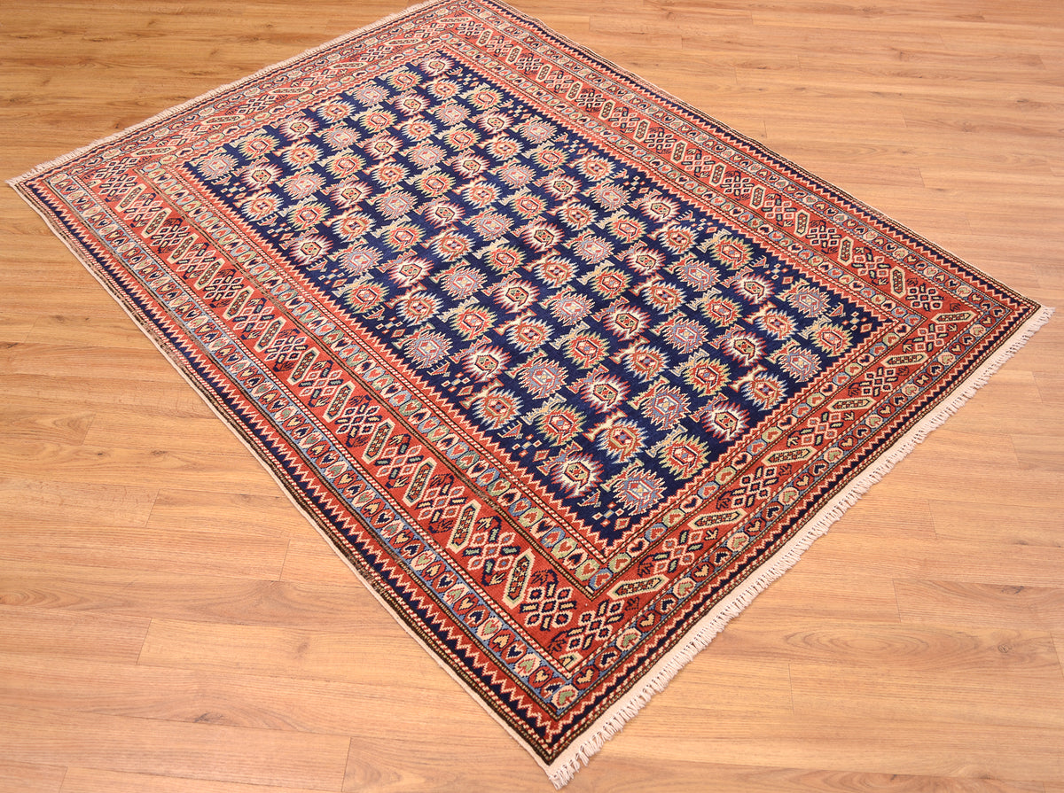 A traditional handmade fine Afghan Shirvan area rug with a navy blue field, rust red borders, and cream motifs, shown laid out on a light brown wooden floor.