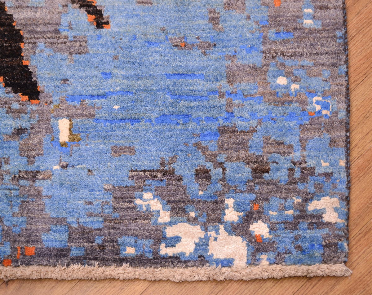 A close-up of a modern patterned handmade carpet with a blue, grey, and beige abstract design, shown on a light wood floor.
