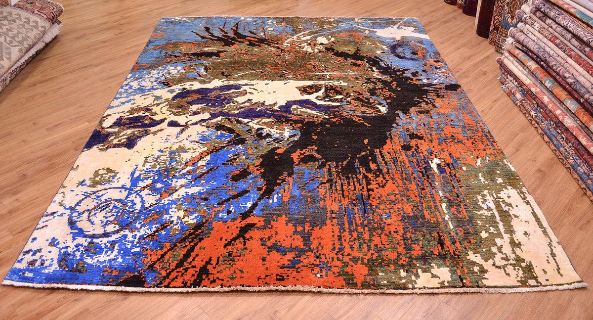 A 10x8 area rug with an abstract, painterly splash design in blue, orange, black, beige, and green, displayed on a wooden floor with stacks of folded rugs visible on the sides.