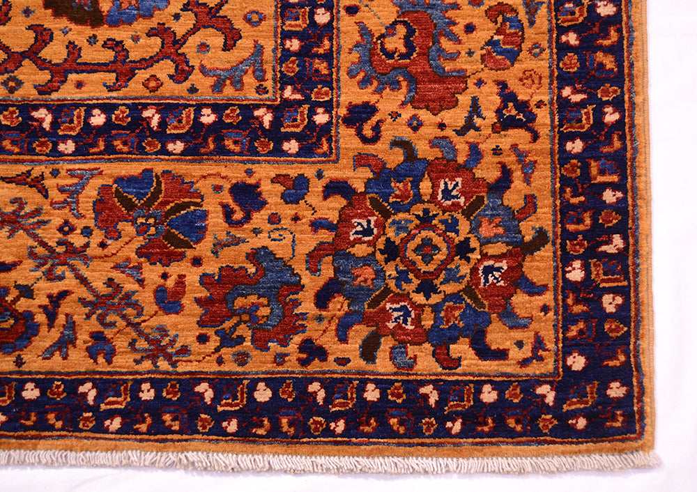 Close-up of a corner of a very fine handmade Suzani rug showing a warm amber field with navy blue borders and red, beige, and blue floral motifs, finished with cream fringe.