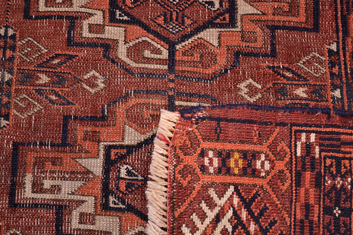 Close-up of a corner turned up of a hand-knotted antique Tekke woven wool rug showing a geometric pattern in rust red, black, beige, and brown, with cream fringe visible at the bottom.