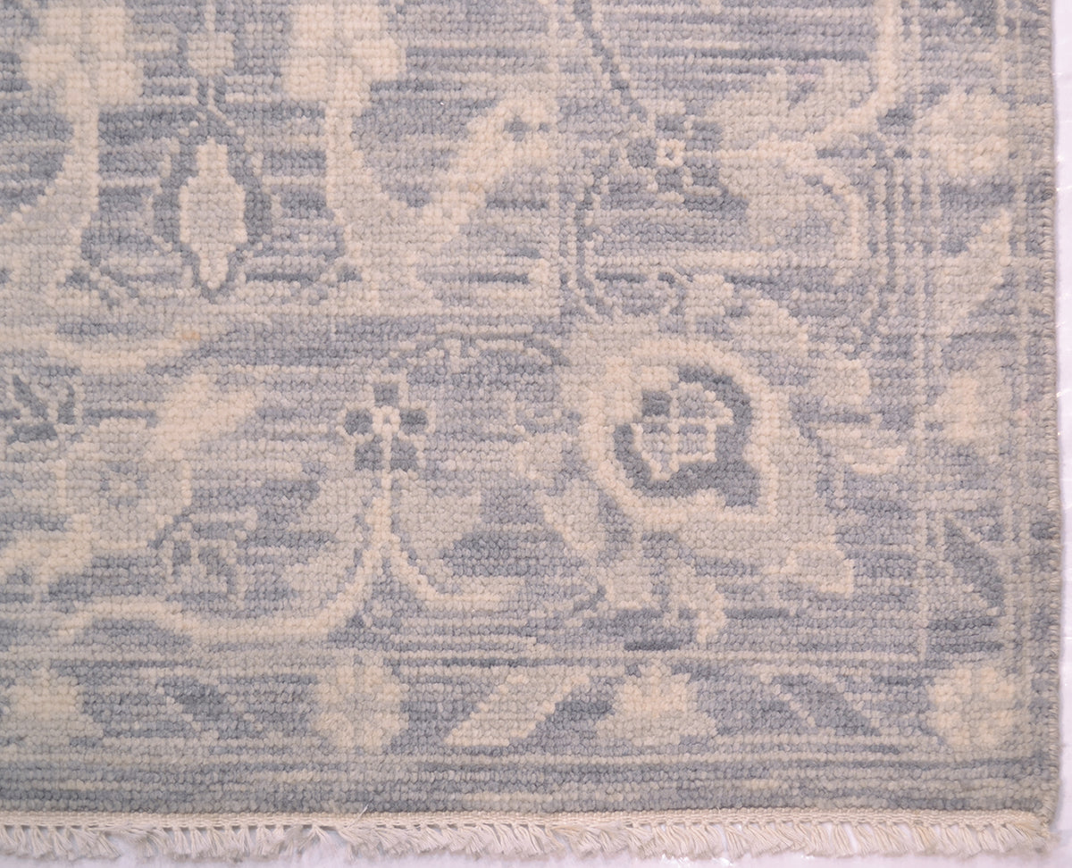 A close-up of a woven tonal rug showing its detailed pattern and texture. The rug features a muted, low-contrast design in dusty blue-gray and warm cream, with a fringed edge visible at the bottom.