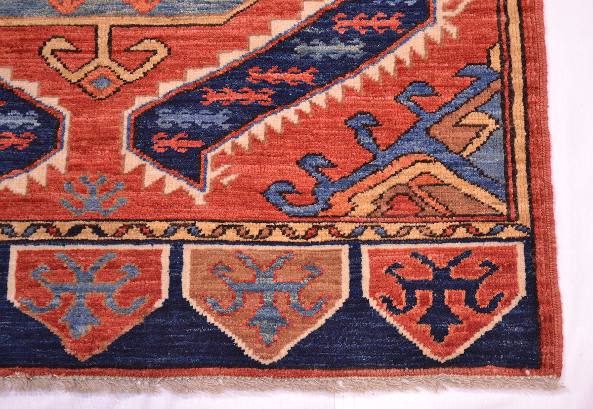 A close-up of an Afghan woven rug corner showing a geometric pattern with rust red, navy blue, beige, and light blue, finished with a fringed edge.
