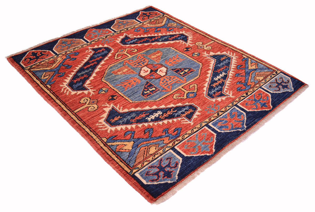 A traditional geometric area square rug with a red-orange field, navy blue borders, and a central blue octagonal medallion, shown from an angled overhead view on a white background.