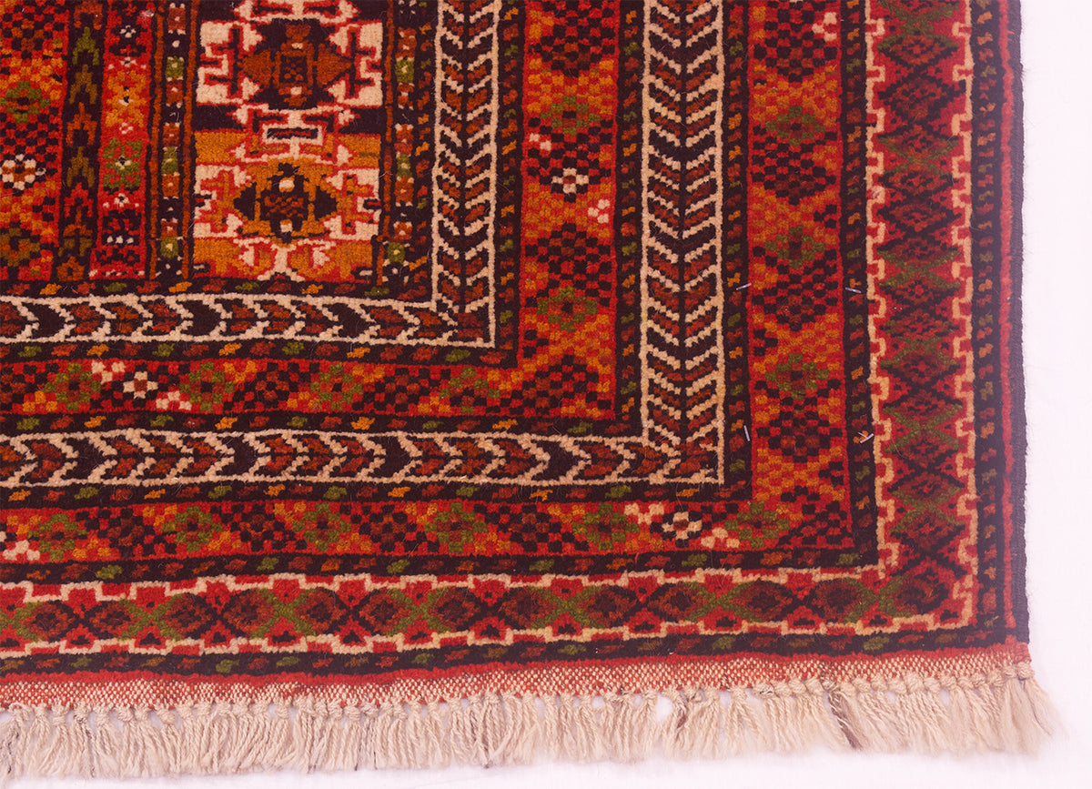 A close-up of a patterned Turkoman rug corner showing geometric borders in rust red, black, beige, and muted green, with cream fringe along the bottom edge.