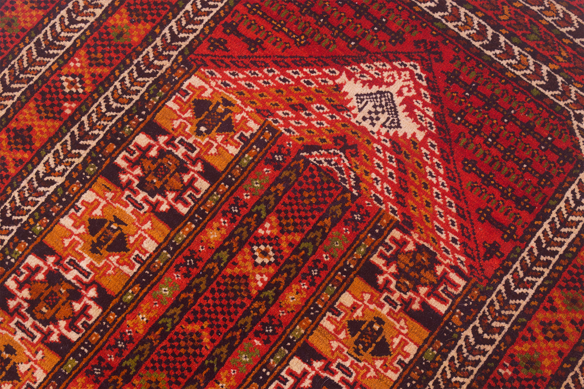 A close-up of a patterned Oriental prayer rug with diagonal bands and geometric motifs in red, orange, brown, black, and cream.