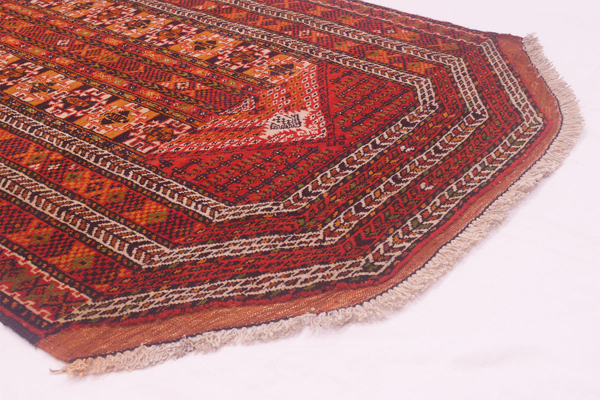 A close-up of a traditional patterned Unusual Turkoman Prayer rug corner showing a complex geometric design in red, orange, brown, and beige with fringed edges on a white background.