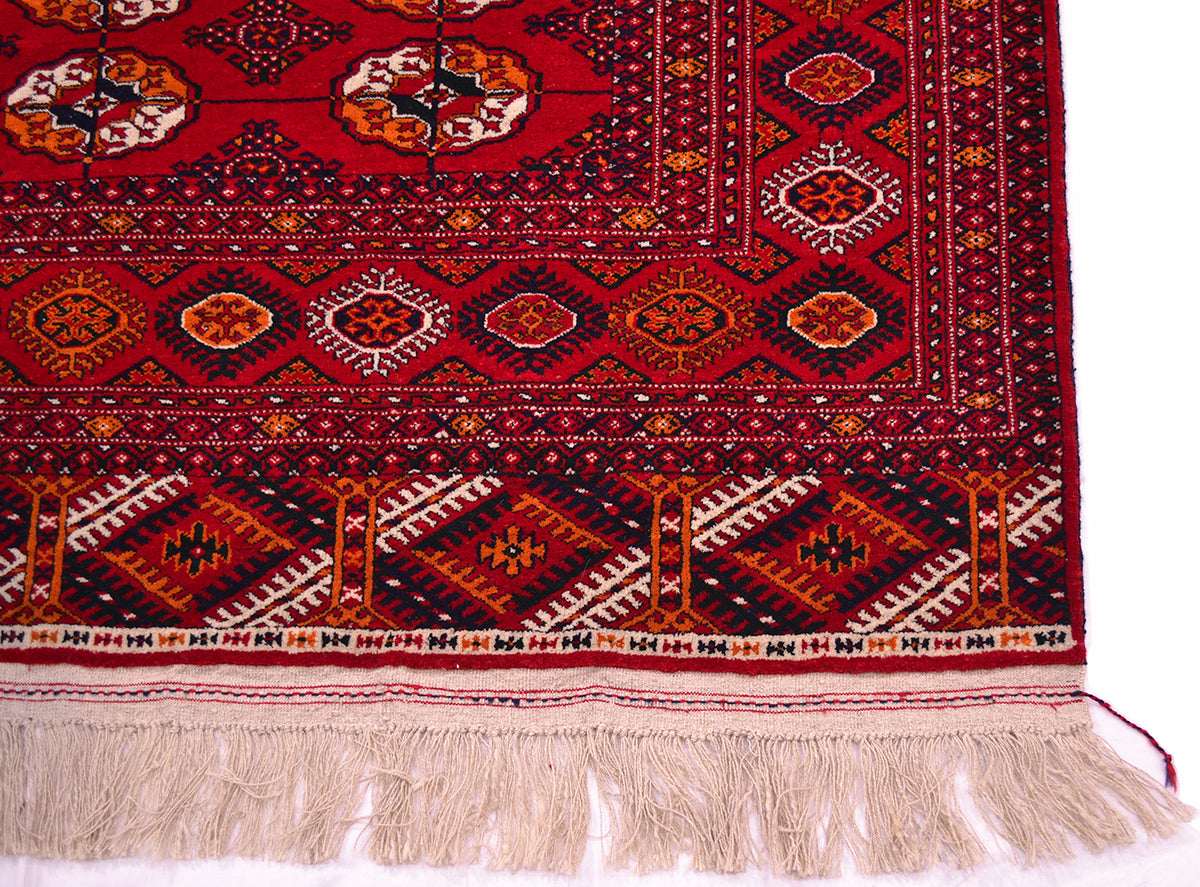 A close-up of a patterned Bokhara Turkoman rug corner showing a deep red field with geometric and floral motifs in black, beige, orange, and white, finished with a cream-coloured fringe.