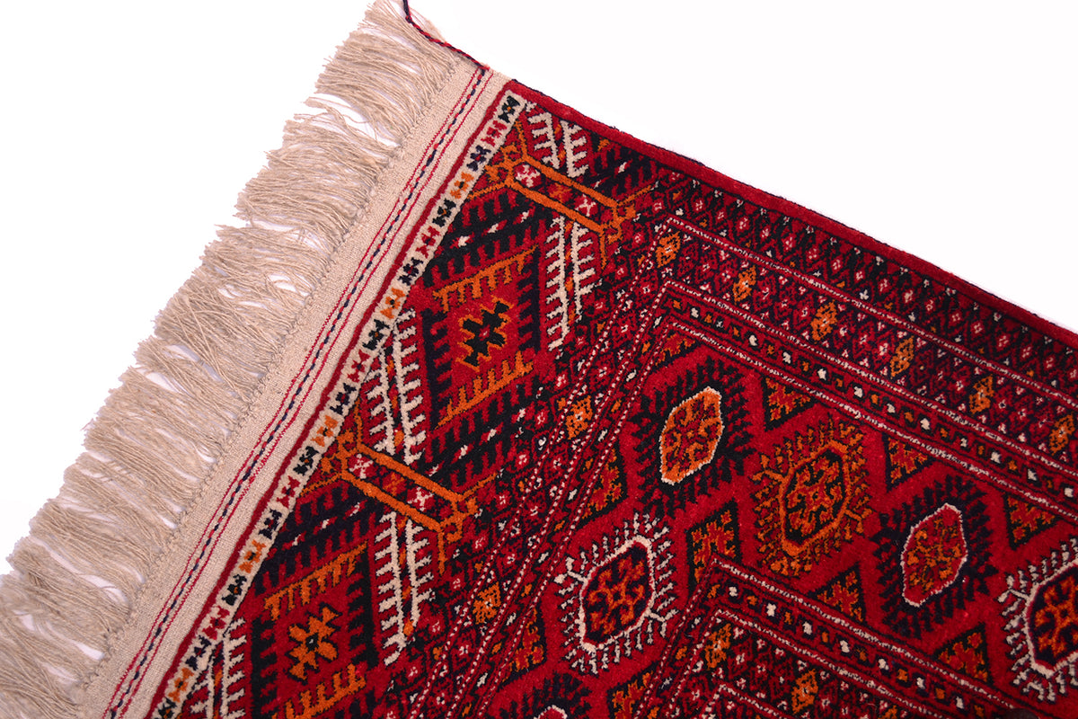 A close-up corner view of a patterned handmade Turkoman red Bokhara rug with cream fringe, featuring a deep red field with geometric and floral motifs in black, orange, and cream on a white background.