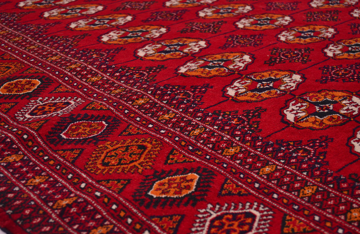 A close-up of a red patterned Bokhara Turkoman rug with geometric borders and floral motifs in orange, cream, and dark blue.