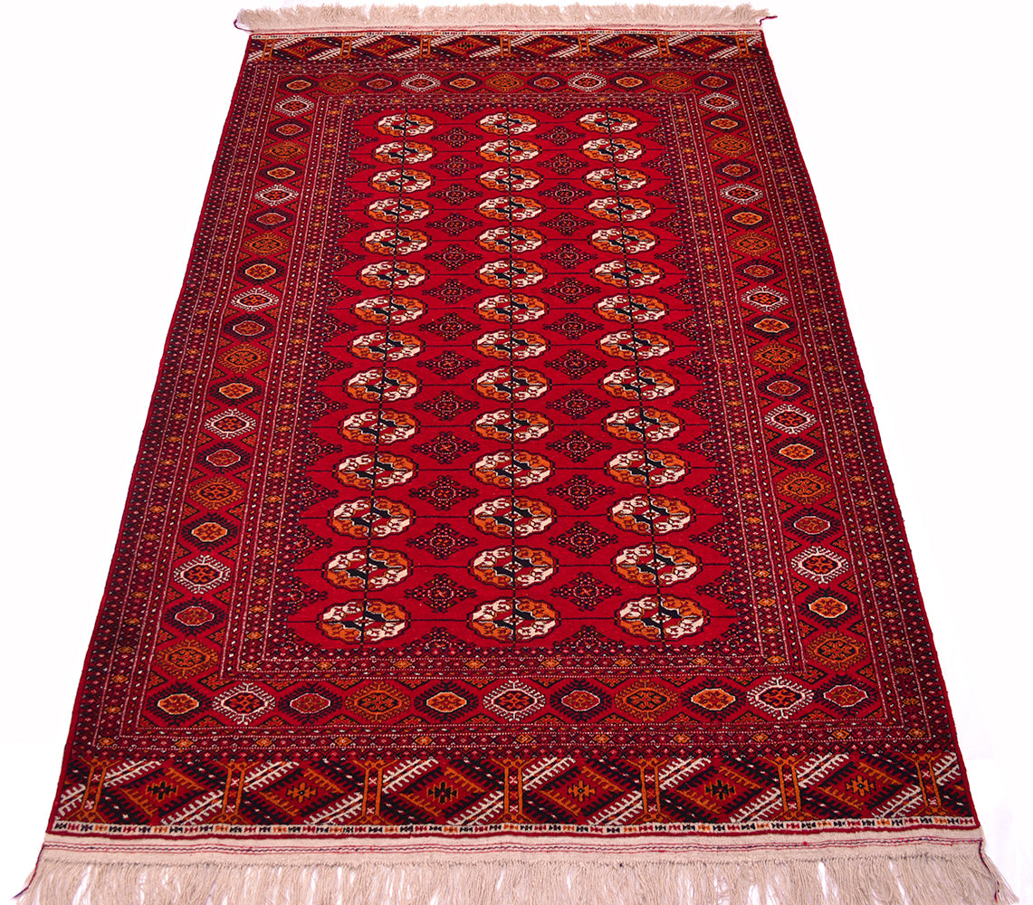 A traditional red Turkoman Bokhara wool rug with a central field of repeating Tekke gul motifs, bordered by ornate geometric and floral patterns, and finished with cream-coloured fringe on both ends.