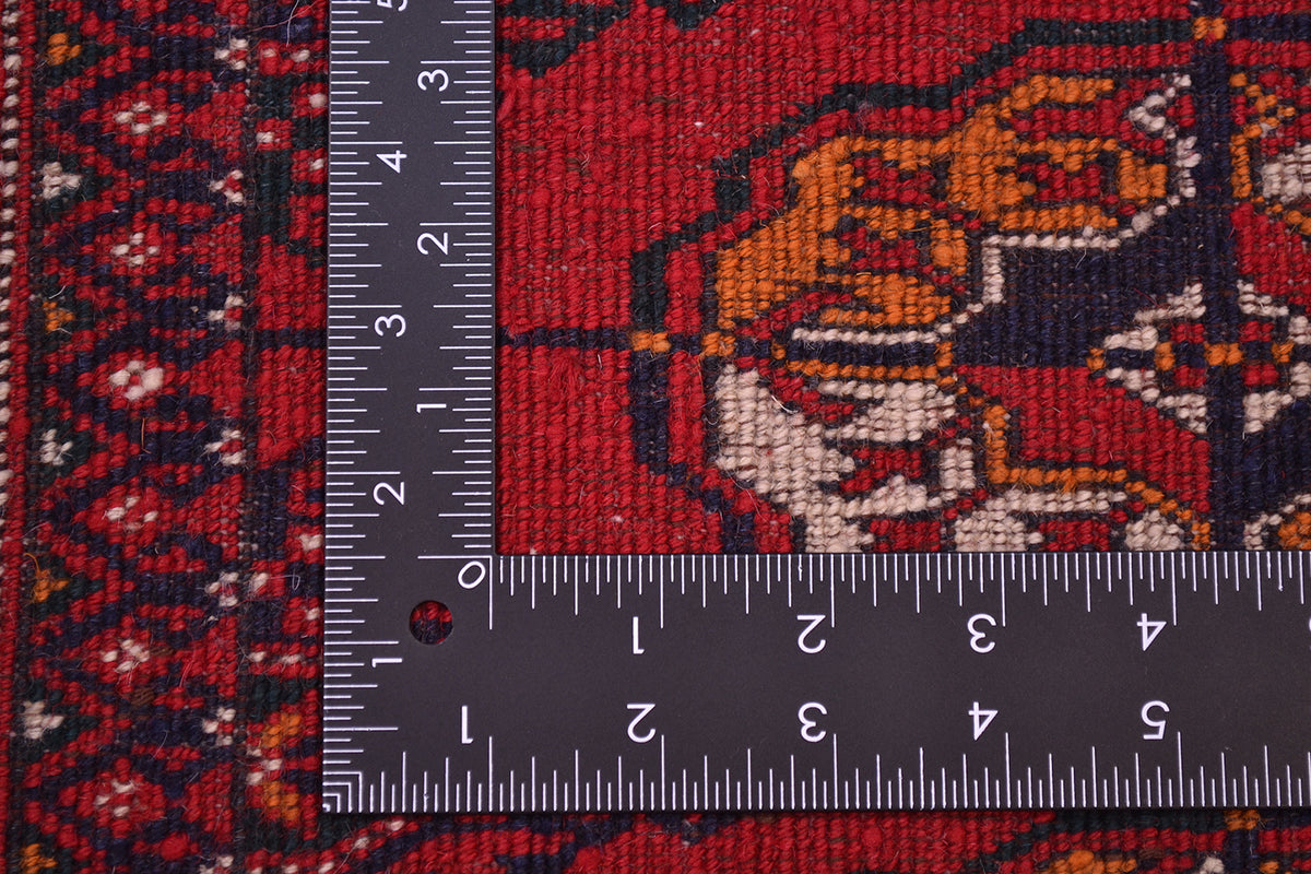A close-up of the hand-knotted weave of a patterned Turkoman Bokhara red rug with a dark metal L-square ruler placed along the left and bottom edges to show scale of the knot density. The rug has a deep red field with orange, cream, and dark navy geometric and floral motifs.