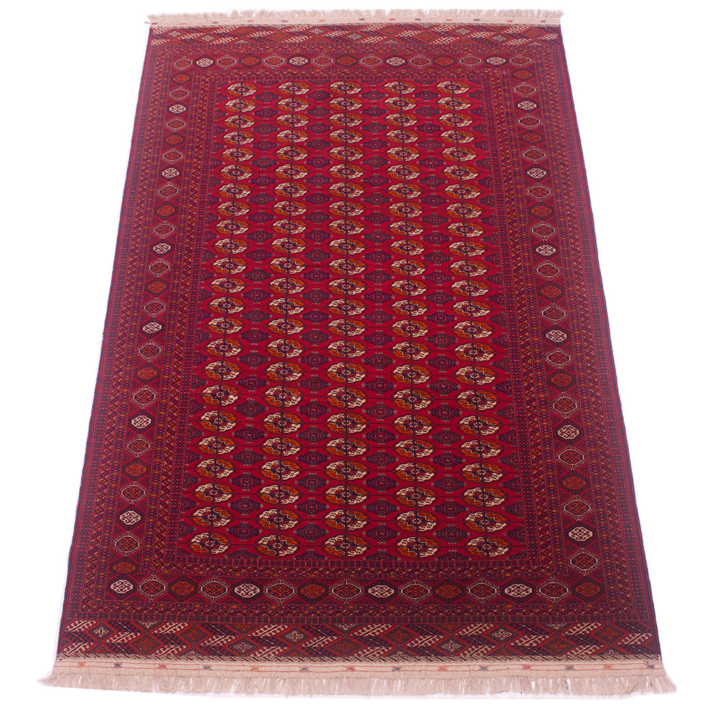 A traditional rectangular Turkoman Bokhara Carpet with a deep red field, featuring vertical columns of repeating medallion motifs in beige, brown, and blue, surrounded by a detailed patterned border and cream fringe on the ends.