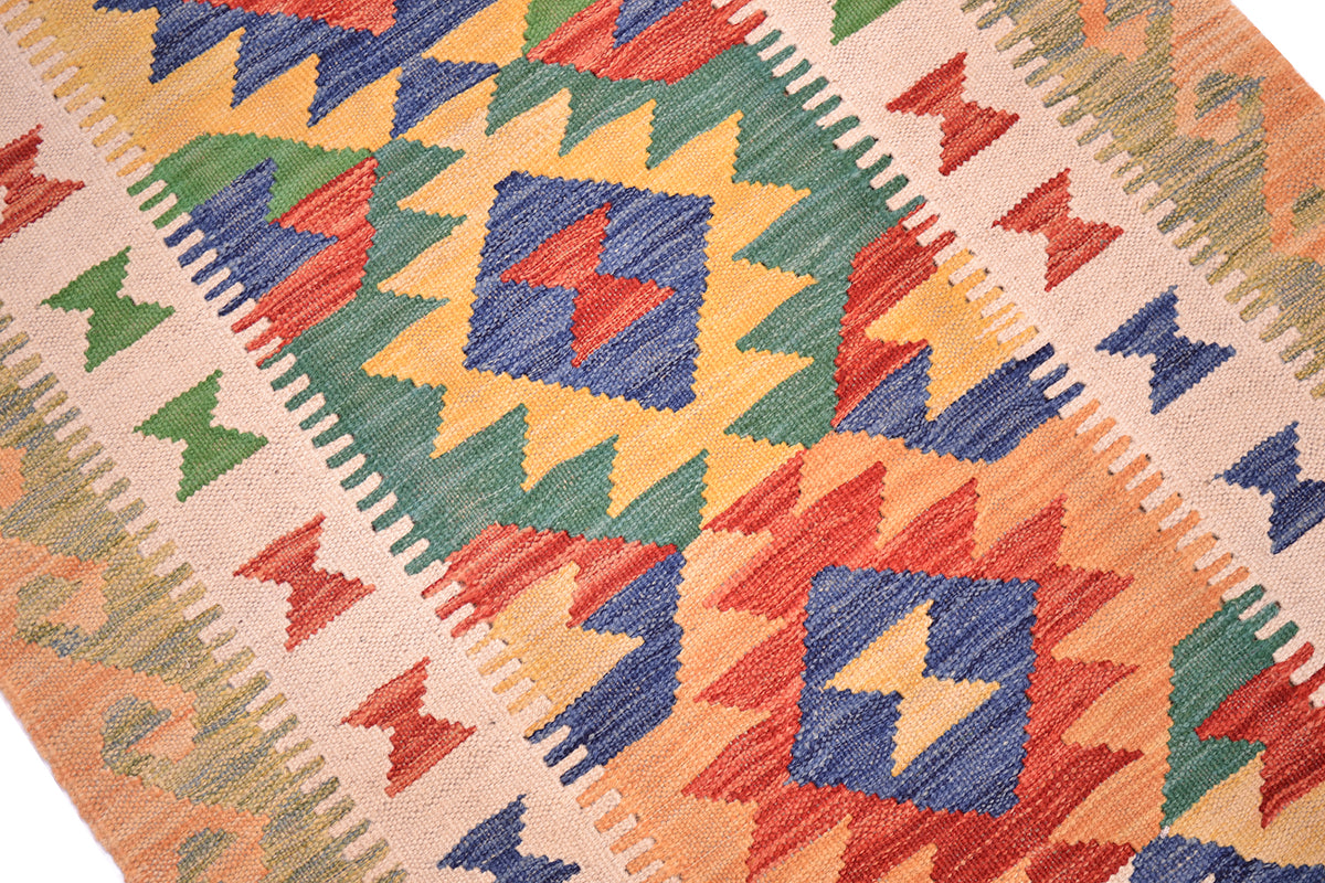 A close-up of a woven kilim rug featuring a vibrant geometric pattern with orange, blue, green, and red shapes on a beige background.