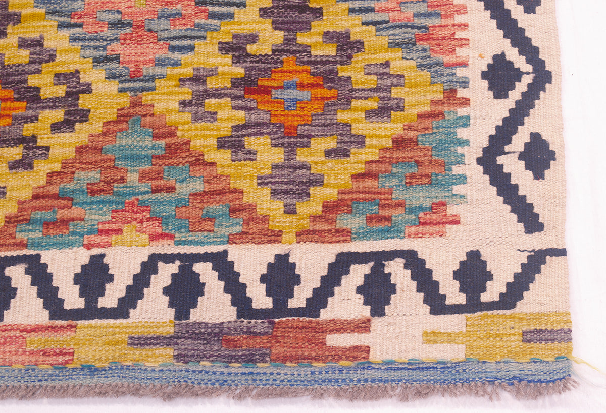 A close-up of a corner of a handwoven Afghan kilim rug showing its geometric pattern and fringed edge against a white background.