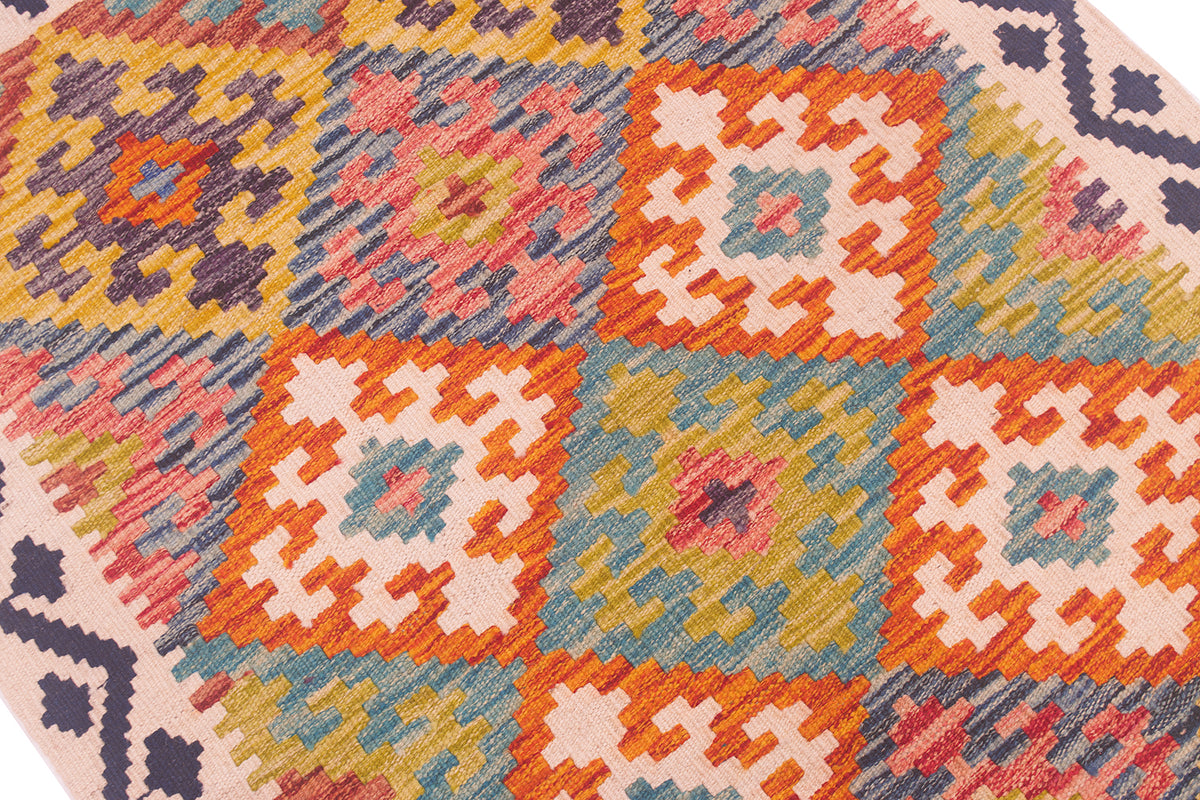 A close-up of a woven kilim rug featuring a colourful geometric patchwork design with orange, pink, yellow, blue, and beige squares.