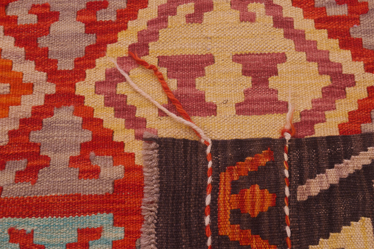 Close-up of a woven red Afghan kilim rug showing a geometric pattern in red, orange, beige, and brown with visible loose threads.