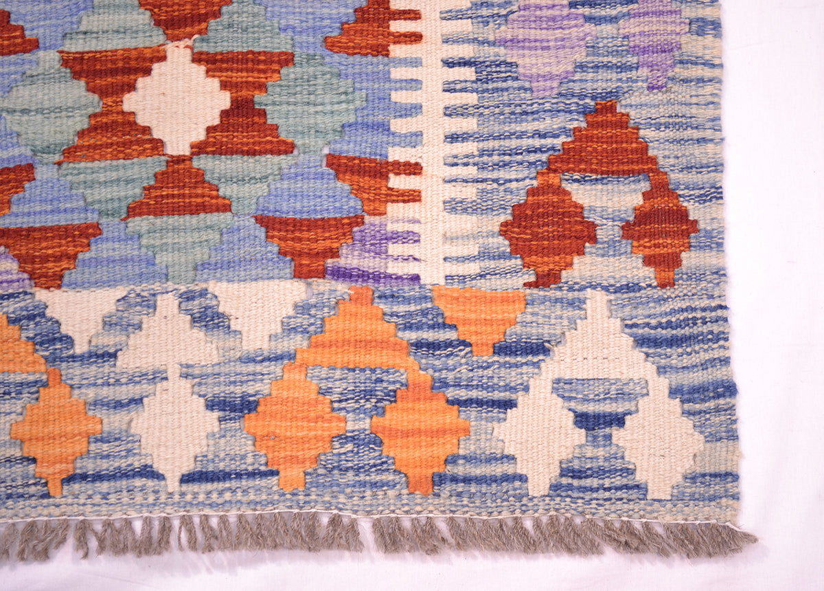 A close-up of a corner of a woven kilim runner showing geometric diamond and triangle patterns in blue, rust red, orange, cream, and light purple, with a fringed edge.